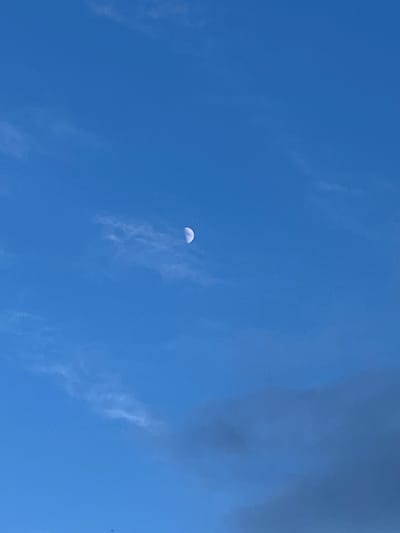 This media file shows a bright blue sky with scattered, wispy clouds. In the center-left of the frame, a crescent moon is visible, illuminated by sunlight. The moon appears as a thin, curved sliver against the vast expanse of the sky. The clouds are thin and translucent, allowing the blue of the sky to be seen through them. A larger, darker cloud formation is present in the upper right corner of the image, partially obscuring the sky. The lighting suggests it is daytime, likely mid-morning or afternoon, given the brightness of the sky and the visibility of the moon. The scene is a natural outdoor setting, with no specific location cues other than the sky itself. No people or specific objects are present in the frame. The overall mood is serene and expansive.