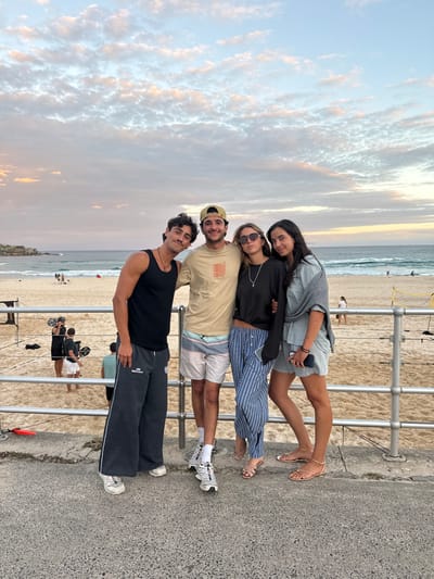 Four young adults are photographed standing on a grey concrete promenade at Bondi Beach, Australia, during twilight. A light grey metal railing, composed of three horizontal bars and vertical posts, is positioned directly behind them, separating the promenade from the sandy beach and ocean beyond. The sky above features scattered cirrocumulus clouds illuminated with hues of light blue, pink, and orange.

From left to right, the individuals are:
1.  A male, estimated 20s, with dark hair and a mustache, wearing a black sleeveless tank top, dark grey wide-leg track pants with white side stripes, and white athletic shoes. His right arm rests on the shoulder of the male beside him.
2.  A male, estimated 20s, with dark hair and a beard, wearing a beige short-sleeve t-shirt, a backward beige baseball cap, patterned board shorts, and white athletic shoes with socks. His right arm is around the shoulders of the female beside him.
3.  A female, estimated 20s, with long dark blonde hair and dark sunglasses, wearing a black long-sleeve crop top, blue and white striped wide-leg pants, and light-colored flat sandals. Multiple silver necklaces are visible. Her left arm is around the waist of the female beside her.
4.  A female, estimated 20s, with long dark hair, wearing a light grey short-sleeve top, a dark grey sweater draped over her shoulders, light grey shorts, and gold-colored flat sandals. She holds a black smartphone in her left hand.

On the sandy beach in the midground, several human figures are present. To the left, a group of individuals surrounds photography or filming equipment, including a large circular reflector and tripods. Further right, a yellow volleyball net is visible. The Pacific Ocean in the background displays light surf breaking near the shoreline. A rocky headland is visible in the far left background. The ambient lighting indicates late evening.