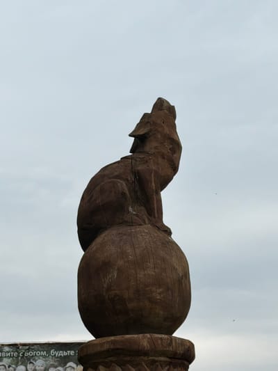 A carved wooden sculpture of a wolf howling stands atop a sphere, positioned against a cloudy sky. The sculpture is dark brown and appears weathered. Below and to the left of the sculpture, a banner with text and images of people is visible. The text on the banner is in Russian and reads "Живите с Богом, будьте!". This sculpture is located in Srednie Kichi, Russia.