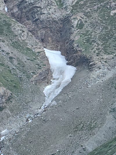 The image depicts a high-altitude mountainous landscape with rocky terrain and patches of green vegetation. A prominent feature is a large snowdrift or glacier situated in a ravine, with a small stream flowing from its base. The snow is a bright white, contrasting with the earthy tones of the surrounding rocks and slopes. The overall impression is of a rugged, natural environment. The location is identified as Kead, India, a region known for its mountainous terrain.