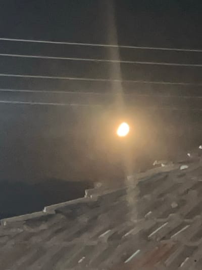The image shows a glowing light source, likely a streetlamp or the moon, obscured by what appears to be a building facade or architectural feature. The structure is composed of repeating linear elements, possibly metal or concrete, creating a textured pattern against the dark sky. To the left of the light source are vertical lines, possibly wires or cables. The location in Kribul, Bulgaria is not evident from the visual information provided in the image.