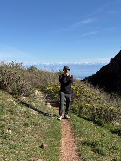 The image shows a person standing on a narrow, dirt path in a mountainous area. The person is wearing a black jacket, black pants, and a hat. They are holding a phone up, presumably taking a picture. To the left of the path is a mountain range, partially visible, with a clear, blue sky above. The right side of the path consists of vegetation, including green grass and bushes, some with yellow flowers. The scene is likely located in the vicinity of Cholpon Ata, Kyrgyzstan, considering the terrain and possible vegetation.