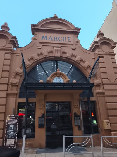 The image shows the exterior of a building labeled "MARCHÉ" in Beausoleil, France. The building has an ornate, arched entrance with a large glass window and decorative elements on the facade. A portrait of a man is visible above the entrance. To the left, there is a large informational sign with historical images and text. The entrance area appears to be a marketplace or retail space, with glass doors and windows revealing the interior. A white metal railing is in the foreground, and a yellow mailbox is mounted on the right side of the building. The sky is a light blue, indicating it is daytime.