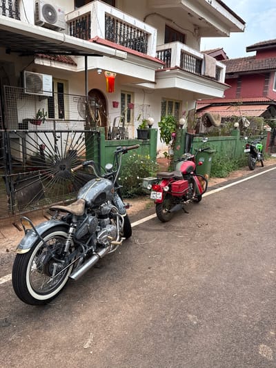The image depicts a street scene in what appears to be a residential area. Three motorcycles are parked along the side of the road. The closest motorcycle is gray with chrome accents and white wall tires. Behind it is a red and green motorcycle. Further down the road is a green motorcycle. Buildings flank the street, featuring various architectural styles and colors. Some buildings have decorative balconies and air conditioning units. A metal fence separates the buildings from the street. Vegetation is visible alongside the fence and between the motorcycles. The road surface is asphalt with a white dividing line. The sky is partially cloudy.