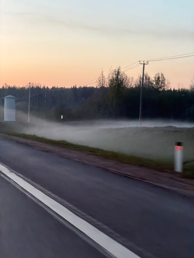 The image shows a scene near Skotnoe, Russia, with a road stretching into the distance alongside a forest edge obscured by fog or mist. The sky displays a gradient from light orange at the horizon to pale gray above. Power lines run parallel to the road and forest. A water tower is visible in the distance. A road sign is present on the right side of the road.