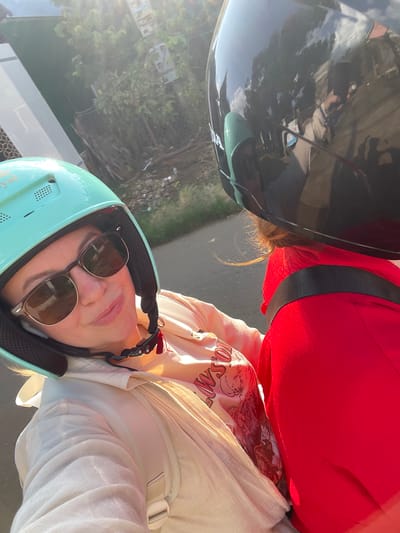 Two individuals are depicted riding a motorbike in Thôn 4, Vietnam. The person in the foreground, facing the camera, is a female wearing a light teal open-face helmet with "Royal" text visible and dark-lensed sunglasses with a brown frame. Her mouth is closed with a slight upward curve. She is wearing a light-colored, long-sleeved outer garment over a t-shirt displaying a graphic design with partially visible text "WSON" and imagery of mountains and animals. A light-colored backpack is worn over her outer garment. The person seated in front of her, facing away from the camera, is wearing a glossy black full-face helmet with "HONDA" visible on the side. The black helmet reflects a cloudy blue sky, trees, and a partial reflection of the individual holding the camera. This person is wearing a bright red long-sleeved garment, with a dark strap (possibly from a backpack or safety restraint) extending horizontally across their upper back. Light-colored hair is visible beneath the back edge of their helmet, illuminated by direct sunlight. The scene is outdoors under strong sunlight, casting highlights and some lens flare from the upper left. The background features a paved road, green foliage, trees, and a light-colored building with a patterned section on the left.