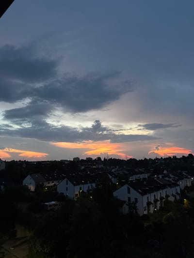 The image captures a twilight sky over a residential area in Sindelfingen, Germany. The sky is dramatic, with dark, heavy clouds in the upper portion transitioning to a band of orange and yellow hues near the horizon, indicating sunset or sunrise. Below the sky, a dense line of trees separates the horizon from a cluster of white, terraced houses with dark roofs. Some houses have illuminated windows, suggesting indoor activity. Streetlights are visible, casting a soft glow on the surrounding greenery and house facades. In the foreground, the scene becomes darker, with more trees obscuring the view of the houses and a glimpse of a caravan parked amongst the foliage. The overall impression is of a quiet evening in a suburban setting.