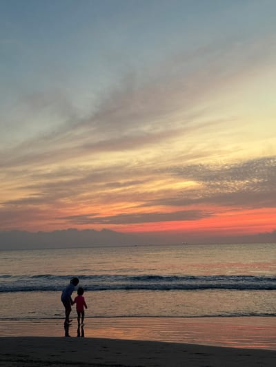 A vibrant sunset fills the sky with orange, yellow, and gray clouds. The ocean reflects the sunset's colors, meeting the sandy beach. Two silhouetted figures, possibly a parent and child, stand at the water's edge, their reflections stretching across the wet sand. The scene evokes a sense of peace and togetherness against the backdrop of a beautiful natural display.