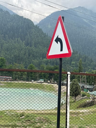 A traffic sign with a black left-turn arrow on a white background, bordered in red, is in the foreground. Behind the sign, a rusty metal fence stretches across the frame. Beyond the fence, a body of light blue water is visible, bordered by green grass. In the background, a dense forest covers rolling hills, leading to mist-shrouded mountains under a cloudy sky. Several vehicles, including a bus and cars, are parked on a road running parallel to the water. The scene is set in Kullan, India.