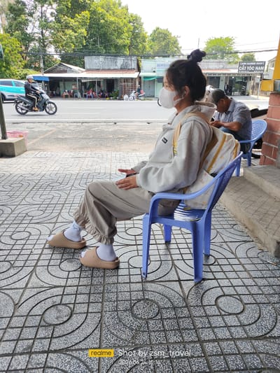 A young woman wearing a white face mask and a grey hoodie is sitting on a blue plastic chair on a sidewalk in Gò Dầu, Vietnam. She has her hair tied up in a bun and is carrying a backpack with a duck pattern. She is wearing beige sweatpants and beige slide sandals with white socks. To her right, an older man in a blue checkered shirt is sitting on another blue plastic chair. In the background, there is a street with a turquoise car and a person riding a motorcycle. Several buildings are visible along the street, including a barber shop and a showroom. The sidewalk is paved with grey and white tiles arranged in a decorative pattern. The timestamp "2026.06.24 10:14" is visible in the lower right corner of the image.