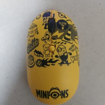 This image, taken in Gimpo-si, South Korea, depicts a yellow, oval-shaped wireless computer mouse positioned on a light gray, textured surface. The mouse features a cluttered, black graphic design primarily consisting of characters and motifs from the "Minions" franchise. Prominent illustrations include various Minion characters in different poses, a Minion skateboarding, a megaphone, a traffic cone, a teddy bear, and the text "Bello." At the bottom front, the word "MINIONS" is printed in black, with the "O" stylized as a Minion's goggle-eye. The mouse has a dark blue scroll wheel centrally located near the top, with a small orange ring around its axis. A faint reddish-brown mark is visible on the mouse surface near the top edge of the scroll wheel. No people or specific interactions are visible. The environment appears to be an indoor setting with diffuse lighting.