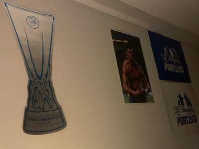 A wall displays a collection of sports-related items. On the left is a large sticker of the UEFA Europa League trophy with "FC Porto x Manchester Utd 31.10.2021" printed on it. To the right is a poster of a muscular soccer player. Further right are two banners, one blue and one white, both bearing the words "La Familia Portista" and an image of a jersey. The lighting is dim, casting shadows across the wall.