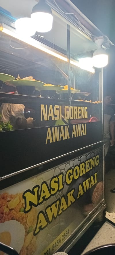 This nighttime image captures a street food stall, likely in the city of Rhieng Blang, Indonesia, illuminated by multiple bright overhead LED light fixtures. The primary subject is a food cart with prominent signage. The upper part of the cart, behind a clear glass panel, displays several light green plastic plates holding various fried food items, possibly fish, chicken, or breaded snacks, resting on black bases. Below this display, a black panel features the text "NASI GORENG AWAK AWAI" in large yellow capital letters. The lower section of the cart's side panel is a graphic advertisement also featuring "NASI GORENG AWAK AWAI" in yellow text with a black outline, overlaid on an image of fried rice with a fried egg and a patterned background resembling stone or tile. A phone number, "221 8454 2684", is visible at the bottom of this graphic.

On the left side, behind the glass and the upper sign, a partial reflection of a person in a light-colored, long-sleeved garment is discernible. On the far right, partially visible behind the cart's frame, is a person wearing a two-toned shirt (light on top, dark below), standing adjacent to the stall. The environment is dark, suggesting an outdoor setting at night, with the ground appearing to be concrete or asphalt.