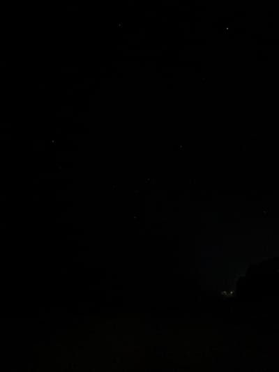The image depicts a nighttime scene over La Cruz de Huanacaxtle, Mexico, characterized by extreme darkness and minimal illumination. The upper two-thirds of the frame show a black sky dotted with numerous faint white points of light, identified as stars. The lower right portion reveals a dark, indistinct silhouette, possibly a distant landmass or vegetation, against the horizon. Within or behind this silhouette, a cluster of a few very dim, yellowish-white artificial lights is visible, indicating distant human presence or infrastructure. No identifiable people, explicit actions, or specific objects beyond celestial bodies and distant light sources are discernible. The environment is outdoors, under a clear, unlit night sky, consistent with a low-light photographic capture.
