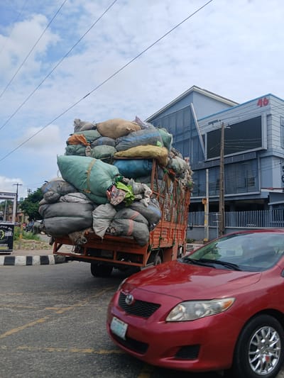 In Animashawun, Nigeria, a large truck is heavily loaded with numerous large sacks, appearing to be filled with various materials. The truck is red with an orange flatbed, and the sacks are tied down with ropes. Some sacks are green, while others are beige, gray, and blue. The truck is positioned on a road, with a red car in the foreground also on the road. The car is a sedan with a license plate that is partially visible. In the background, there is a modern building with a sign that reads "46". The sky is cloudy. There are utility poles and wires visible, as well as some trees and a street sign.