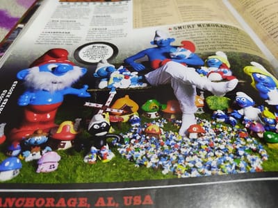 The image displays an open book, presenting a detailed arrangement of Smurf figurines and related miniature objects on a simulated green, grassy surface. A large Papa Smurf figure, with a red hat and white beard, stands prominently on the left. To its right, an unclothed blue Smurf figure and another Smurf figure with a red hat are seated on a brown bench-like object. A human leg, clothed in white socks and light-colored trousers, extends from the upper right, with the foot resting on the miniature grass. The scene includes various other Smurf figures, miniature colorful mushroom houses, a small windmill, and hundreds of tiny, multi-colored Smurf-like figurines densely scattered across the ground. Text on the visible page sections mentions "SMURF MEMORABILIA" and "Anchorage, AL, USA". No information within the image specifies the location as Harasta, Syria.