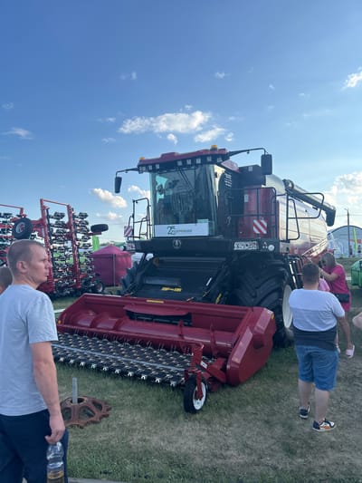An outdoor agricultural exhibition is underway in Prutskoy, Russia. The primary focus is a large, red combine harvester with a wide cutting platform. Several people are present, observing the machinery. To the left of the combine, a man in a gray t-shirt and dark pants stands holding a bottle of liquid. In the background, other agricultural equipment, including what appears to be a planter or seeder with numerous tines, is visible. Tents, one purple and one green, are also set up at the exhibition. The sky above is blue with scattered white clouds. The ground is covered in grass.