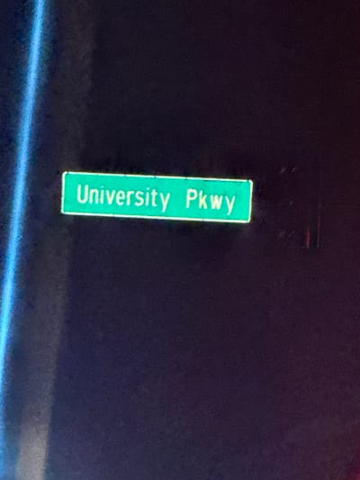 A vertical street sign reads "University Pkwy" in light green lettering against a dark green background. The sign is illuminated, standing out against the dark surrounding area. The image quality is somewhat grainy, particularly in the darker areas.