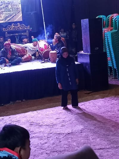 This media file captures a nighttime outdoor cultural event in Pangalengan, Indonesia, illuminated by artificial blue and purple lighting. The scene features a raised stage where several male musicians are performing, and a woman stands in front of the stage, observed by an unseen audience in the foreground.
On the stage, multiple men are actively engaged in music. Two men are prominently playing large traditional Indonesian drums (kendang or gendang). One musician on the left wears a red head covering, while another in the center sports a white long-sleeve shirt and a green beanie. A man on the right side of the stage wears a traditional Indonesian headpiece (iket or blangkon). Other figures are visible in the background on and around the stage, suggesting additional performers or crew. Small plastic cups are placed on the stage, possibly containing beverages.
In front of the stage, a woman stands barefoot, clad in a blue tunic, black pants, and a dark headscarf (hijab). She appears to be looking downwards, casting a long shadow on the uneven ground, which suggests dirt or a similar natural surface. In the immediate foreground, the back of an audience member's head is visible.
To the right of the stage, large black speaker boxes are stacked, indicating sound amplification for the event. Beside them, green and orange plastic chairs are neatly stacked, suggesting temporary seating arrangements for attendees. Above the stage, a banner or backdrop displays partially visible text, including numbered items: "7 SINGA DEPOK", "8 WAYANG DAKWAH", and "9 SOUND SYSTEM," likely referring to acts or services at the event.
The overall atmosphere indicates a local community or cultural performance, possibly a traditional music concert or a Wayang Dakwah (shadow puppet show with Islamic preaching) as suggested by the banner text.