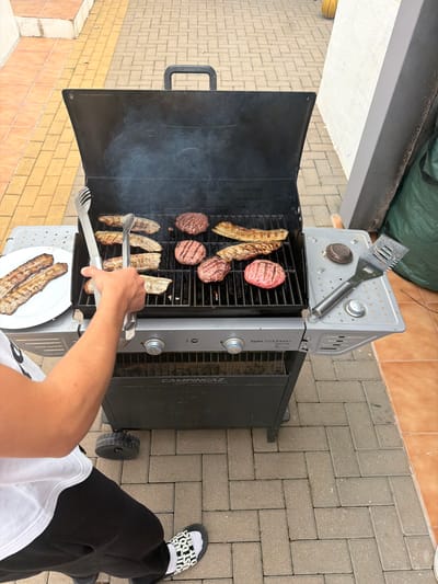 A person is grilling meat on a portable gas grill, outdoors on a brick patio, possibly in Montgó, Spain. The grill has a black lid and a silver top with a grill brush resting on it. Burgers and grilled vegetables are visible on the grill, with some already cooked on a white plate. The person is using tongs to move the food. The person is wearing black pants and a white t-shirt, with "JUST DO IT" sliders on their feet.