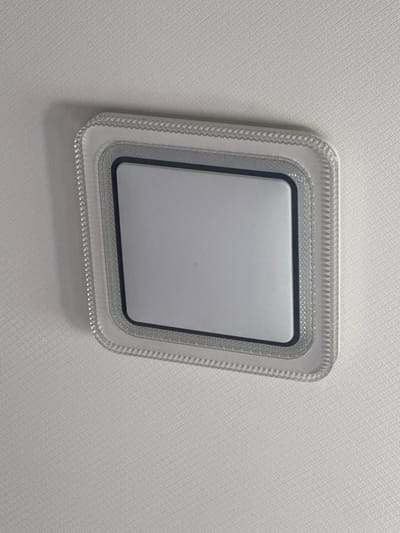 A square ceiling light fixture with a translucent, textured frame and a central, opaque white panel is affixed to a white, textured ceiling. The fixture appears to be centered in the frame. The image was taken from a low angle, looking up at the fixture. The setting is an interior room, likely a residential or commercial space, in the city of Kentau, Kazakhstan. There are no people or visible actions taking place. The environment is clean and well-lit, suggesting a modern or recently maintained interior.