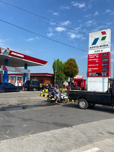 The image shows a gas station in Kecamatan Tulangan, Indonesia, located at the coordinates -7.479 latitude and 112.643 longitude. There are several cars and motorcycles parked on the asphalt lot. Two individuals wearing helmets are riding a white moped on the left side of the image, while a black pickup truck is parked on the right side. The gas station is a Pertamina brand, and the sign displays the prices for different types of fuel, including Pertamax Turbo 98, Pertamax 92, Pertalite, Pertamina Dex, and Dexlite Bio Solar. The sky is blue with white clouds and there are trees surrounding the lot.