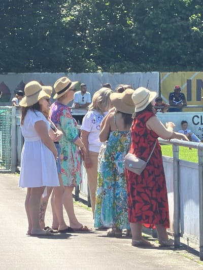 The image depicts a group of five women standing side-by-side, seemingly viewing an event, likely in the city of Fentange, Luxembourg. All are wearing straw hats and dresses. The woman closest to the camera is wearing a white dress, and the next in line is in a light-blue patterned dress. The third woman is wearing a white top and tan pants with a blue-patterned dress, followed by a woman in a blue-floral print dress. The woman furthest from the camera is wearing a red floral-print dress and carrying a small grey purse. They are standing alongside a low metal barrier. People and signage are visible in the background. A dense tree line is on the far left.