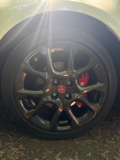 An Abarth wheel with red brake calipers is shown, close-up, in Bouctouche, Canada. The wheel is dark gray and has five spokes, with the Abarth logo visible in red and white at the center. The brake caliper is also red and has a white Brembo logo. The tire is black and has visible tire markings. The background is slightly blurred and shows some foliage on the ground, suggesting an outdoor setting, possibly in a natural or park-like area in Bouctouche. The lighting suggests it's daytime, with sunlight filtering through from the top of the frame, creating lens flare effects.