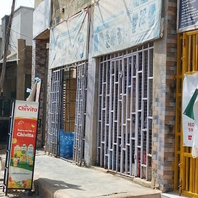 This image depicts a street-level view of a commercial section in Dan Maliki, Nigeria, under bright daylight conditions. The scene features multiple storefronts with visible security gates and informational banners.
On the left, a "Chivita" branded A-frame sidewalk sign, predominantly white and orange, displays images of various juice products and slogans like "Everyone has a Chivita." Adjacent to this, a shop entrance features a white metal security gate, with one panel pulled back, revealing blue tarp-like material or sacks inside the dimly lit interior.
To the right, another shop is secured by a fully closed white metal security gate composed of vertical and horizontal bars. Above this shop, a large, light-colored banner is affixed to the building, displaying text such as "TEXTILE & TAILORING DESIGN" alongside illustrations of garments and patterns.
Further to the right, a bright yellow metal gate, possibly for an additional establishment, is partially visible. Attached to this gate are two banners: a white one with green text and a green graphic, showing "REG" and "SIN," and another white banner with black text listing various services including "DATA SUBSCRIPTION," "NIN REGISTRATION," "BVN REGISTRATION," "ID CARD," "INTERNATIONAL PASSPORT," and "BUY CRYPTO ASSET."
The building facades exhibit a combination of light-colored brickwork or tiling on the lower sections and plain concrete or painted surfaces above. Overhead light fixtures are mounted on the building. The sidewalk in the foreground is paved, and strong shadows are cast, indicating the time of day. No individuals are visible in the frame.