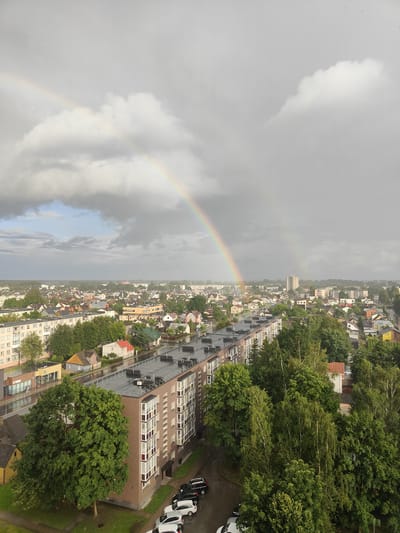This is an aerial shot of a city, likely Panevėžys, Lithuania, under a dramatic sky with a double rainbow. The primary focus is on a long, brown apartment building in the foreground, with a multi-story structure featuring numerous windows and balconies. Several cars are parked in a lot beside the building.
Beyond the apartment building, the landscape transitions to a dense residential area with numerous smaller houses, many with red or orange rooftops, interspersed with lush green trees. A taller, light-colored building can be seen in the distance, suggesting a more commercial or central part of the city. The sky is filled with heavy, grey clouds, but a bright rainbow arcs across the frame, with a fainter secondary rainbow visible above it, indicating recent rain or ongoing showers. The overall atmosphere is moody and ethereal due to the weather conditions and the presence of the rainbow.