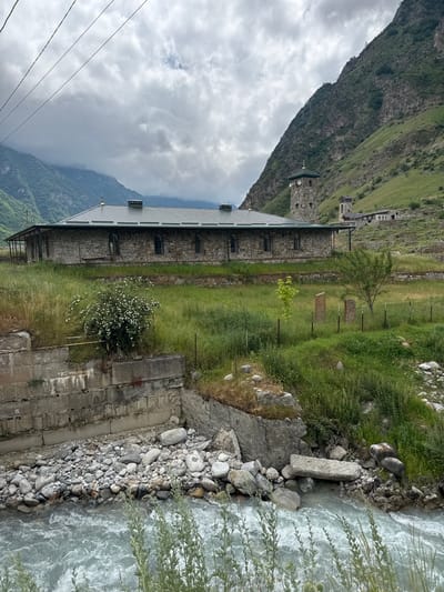 This image depicts a scenic view of a stone building situated beside a turbulent river, with a steep, grassy mountainside in the background. The building appears to be an old, possibly historical structure, constructed with rough-hewn stones and featuring a dark metal roof with several dormer windows. Adjacent to the building, a fence runs along a patch of tall grass and scattered rocks, leading towards the river. The river itself is a powerful, swirling body of water, with many smooth stones lining its banks. In the distance, more stone structures and buildings are visible on the lower slopes of the imposing mountain. The sky is overcast with heavy clouds, suggesting a cool, possibly damp, day. The overall atmosphere is one of natural beauty and historical presence, hinting at a remote and rugged location. The image is captured in a vertical orientation, with the landscape appearing sideways. There is no visible text in the image. The scene is likely set in Verkhnyaya Balkariya, Russia, given the mountainous terrain and architectural style.
