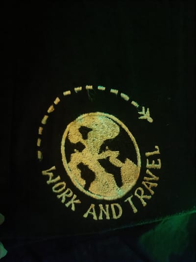 The image shows a close-up of an embroidered patch on a black fabric. The patch features a stylized globe in the center, depicted with continents in black and landmasses in a textured yellow. Surrounding the globe is a dashed circular line, suggesting a flight path. A small airplane icon is visible along this path, indicating travel.
Below the globe, text is embroidered in yellow capital letters, forming an arc. The visible text reads "WORK AND TRAVEL". The embroidery is detailed, with visible threads creating the texture of the globe and the letters. The overall impression is a graphic design related to globetrotting and professional endeavors.
The lighting in the image is dim, with some green and yellow hues, possibly from ambient light sources or reflections. The black fabric appears to be part of an article of clothing or a bag. There are no people visible in the frame. The setting is not clearly defined beyond the implied context of the "WORK AND TRAVEL" theme. No specific time of day, weather, or emotional cues are discernible.