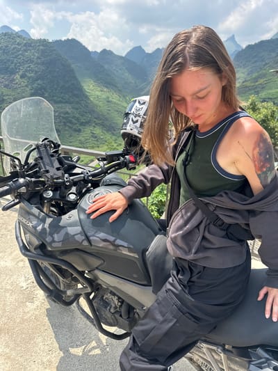 A woman with long brown hair and a tattoo on her left arm is resting on a gray and black camouflage motorcycle in Lũng Phìn Commune, Vietnam. She wears a dark green tank top with white trim, a dark gray hoodie, and dark pants. Her eyes are closed, and she rests her head on the fuel tank with her right hand on top. The motorcycle has a helmet resting on the left handlebar. A clear windscreen is visible. In the background, a mountain range extends into the distance. The sky is partly cloudy.