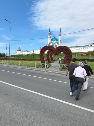 In Усады, Russia, on a bright day with a blue sky and scattered clouds, a wide asphalt road stretches across the foreground. A white dashed line divides the road. In the middle ground, a grassy area is bordered by a metal fence. Dominating this area is a large, red floral sculpture shaped like two intertwined hearts, with a pathway leading through it. Beyond the floral display, a white fortress wall with turrets and towers extends into the background. A prominent blue-domed mosque with two minarets stands against the sky behind the fortress. Four people are walking away from the camera on the road, in the foreground. The person closest to the camera on the left wears a dark t-shirt and dark pants, followed by a person in a light pink shirt and dark pants, then a person in a black top and light pants. A fourth person, mostly out of frame to the right, wears a black top and light pants, carrying a red bag. Several other people are visible walking in the background near the floral sculpture and on the fortress grounds.
