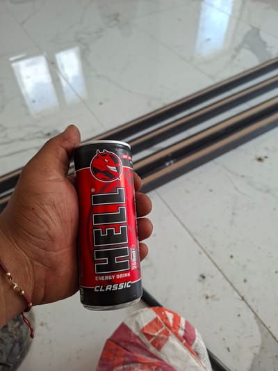 A person's hand is holding a can of "HELL ENERGY DRINK CLASSIC" in the city of Dolna Malina, Bulgaria. The can is red and black with a devil logo. The background is a tiled floor with reflections and dark horizontal bars. A braided bracelet is visible on the person's wrist.