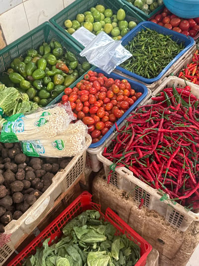 The image shows a close-up view of various fruits and vegetables displayed in baskets at a market stall. A blue plastic basket contains small green chili peppers. Adjacent to it, a white plastic basket is filled with long, red chili peppers. Another blue basket contains small, oval-shaped red tomatoes. Additional green vegetables and fruits are visible in the surrounding baskets. Two packages of Enoki mushrooms in green and white packaging are positioned in front of a basket of dark brown, round vegetables. A red plastic basket in the upper right contains green leafy vegetables. The image, taken in Gunung Putri, Indonesia, focuses on the colorful array of produce available at the market.