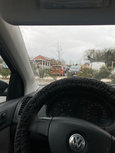 The image depicts a scene observed from the driver's seat of a parked vehicle, specifically in the parking lot of a McDonald's restaurant located at Avenue du Général de Gaulle, 95150 Taverny, France.

In the foreground, the lower portion of a black steering wheel with a fuzzy, dark grey cover is visible. The steering wheel prominently displays a silver Volkswagen logo. To the right of the steering wheel, a portion of the vehicle's instrument cluster is visible, showing a speedometer marked up to 220 km/h, a tachometer, and a digital display indicating "1640" (likely kilometers). The top edge of the car's interior roof and a sun visor are visible at the upper margin of the image. The A-pillar is partially visible on the left.

The midground shows a parking lot filled with various vehicles and landscaping. Directly ahead, a dark orange minivan or SUV is partially obscured by low, green and reddish bushes that separate parking rows. To its right, a dark grey Renault vehicle, possibly a Captur, is parked, displaying the French license plate "DL-236-RN". Further to the right, a white van is visible. Other vehicles are parked in the distance, partially obscured. A distinct, bare deciduous tree stands in the center-left midground.

In the background, a building with a red-tiled roof, white supporting columns, and a covered patio area, characteristic of a McDonald's restaurant, is visible on the left. The sky above is overcast and grey. A line of trees and dense foliage forms the horizon behind the parking lot, some with bare branches and others evergreen. No individuals are distinctly visible or engaged in actions within the frame.