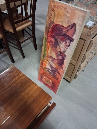 An overhead shot displays a tilted canvas painting of abstract flowers in orange, red, and purple hues. A light brown wooden box sits behind the painting. In the lower left, a dark wooden chair with a woven seat is partially visible. To the right, a polished wooden surface gleams with reflected light. The floor appears to be tiled in a neutral tone.