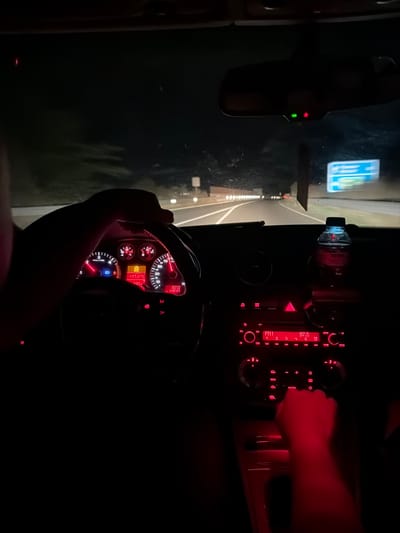 The image shows the interior of a car at night, likely in Belozem, Bulgaria, as indicated by the car's dashboard displaying the location. The driver's hands are visible on the steering wheel, and the dashboard lights, car radio, and surrounding interior are illuminated in red. A bottle of water rests on the dashboard. The car appears to be on a road, with streetlights and other vehicles visible in the distance through the windshield. The overall scene suggests a nighttime drive through a town or city.