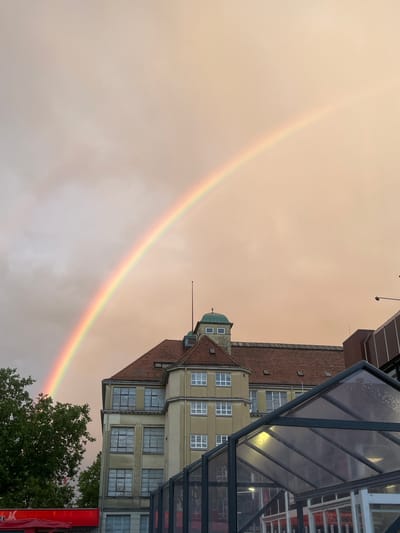 The image depicts an urban landscape under a unique sky, captured after rainfall, which is indicated by droplets on a glass structure in the foreground. The sky is a prominent feature, displaying a warm, hazy orange-brown hue that suggests either a specific time of day like sunset or sunrise, or unusual atmospheric conditions.

A vibrant rainbow forms a gentle arc across the sky, stretching from the lower left to the upper right of the frame (when viewed in correct orientation). Its full spectrum of colors is visible, although slightly softened by the atmospheric haze.

On the right side, several buildings with distinct architectural features are visible. A prominent building in the mid-ground has a reddish-brown tiled roof and light-colored walls with numerous rectangular windows. It features a corner tower-like structure topped with a green dome and additional windows. Another building with a similar tiled roof and windows is situated further back. In the immediate foreground on the far right, a modern structure with a dark metal frame and glass panels, possibly a bus stop shelter, partially frames the scene.

The lush green foliage of a tree is visible in the lower right corner, partially obscuring the base of the rainbow. A red sign, partially hidden behind the glass structure, bears the German text "WOHNTRAUME" (meaning "living dreams" or "home dreams"), with "EL" and possibly "A" visible below it. The scene captures a moment of natural beauty, likely after a rain shower, with the striking contrast of the rainbow against the unusually colored sky.