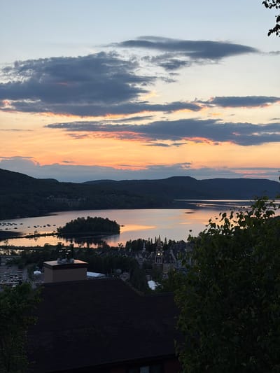 This is a high-angle, medium shot of a lake and a village at sunset in Mont-Tremblant, Quebec, Canada. The sky is a gradient of pale blue at the top, fading to orange and pink hues near the horizon, with scattered clouds. The lake occupies the center of the image, reflecting the colors of the sky. A small, tree-covered island is situated in the middle of the lake. On the far side of the lake, a range of dark, tree-covered mountains forms the horizon.
In the foreground, the roofs of several buildings are visible, some with dark shingles. Among these is a building with a light brown rectangular section topped with multiple metallic chimneys. Further into the scene, nestled between the lake and the mountains, is a village characterized by its distinct architecture, featuring buildings with steeply pitched roofs, dormer windows, and at least one prominent clock tower with a pointed, red roof. Some structures appear to be hotels or residential buildings.
To the left of the village, a parking lot with numerous cars is visible, adjacent to a body of water where several boats are moored. The overall impression is of a serene, picturesque landscape during the golden hour, with the village and natural elements harmoniously coexisting. There are no people or animals visible in the image.