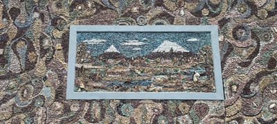 The image captures a mosaic wall in Garni, Armenia. The wall is predominantly composed of small stones, arranged to create a textured, organic pattern. A rectangular mosaic, framed in a plain light gray border, is mounted on the wall. The framed mosaic portrays a landscape scene with two mountains capped with white snow or ice under a blue sky with scattered white clouds. The foreground shows a variety of natural elements like trees and bodies of water. The surrounding wall mosaic is densely patterned with swirling motifs and floral shapes crafted from stones of various earthy tones.