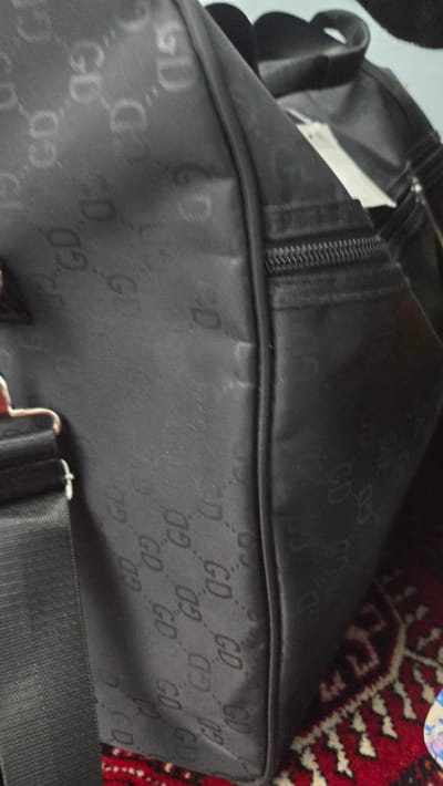 The image shows a close-up, dimly lit view of a black bag with a repeating "GD" logo pattern embossed on its surface. The bag appears to be made of a textured fabric, possibly nylon or canvas. A zipper is visible along the top edge of the bag, and a black strap with silver hardware is partially in view at the bottom. The background features a portion of a rug with a red, black, and white intricate geometric pattern, characteristic of Turkmen carpets. The lighting is low, and the focus is primarily on the bag's texture and logo. There are no people or distinct activities visible, suggesting the image is a product shot or detail shot of the bag, possibly placed on the rug in a domestic setting in Kaka, Turkmenistan. The overall mood is neutral, focusing on the visual details of the object.