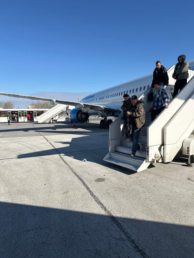 People are deplaning from an airplane at Bolshoye Savino Airport in Bolshoye Savino, Russia. Passengers are descending the stairs from the airplane onto the tarmac. The aircraft is connected to a mobile passenger boarding stair truck, located at the bottom of the stairs. A ground crew person is standing near the truck. The sky is clear and blue.