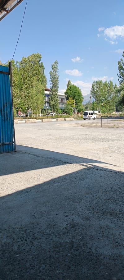 The image depicts an outdoor scene in Yangiariq, Uzbekistan, on a bright, sunny day. In the foreground, a light-colored, gravelly surface is visible, with a distinct shadow cast from the left side of the frame, suggesting an overhead structure or barrier. To the left, a portion of a blue corrugated metal fence is present.
In the mid-ground, a paved area is visible with several concrete barriers. A white van is driving on this paved area. Behind the paved area, a multi-story building with a light-colored facade is situated, surrounded by lush green trees. The building appears to be a hotel or guesthouse, with balconies and windows. Beyond the building and trees, a mountain range can be seen in the distance under a clear blue sky with scattered white clouds. The overall environment suggests a somewhat rural or semi-urban setting with natural elements prominent.
