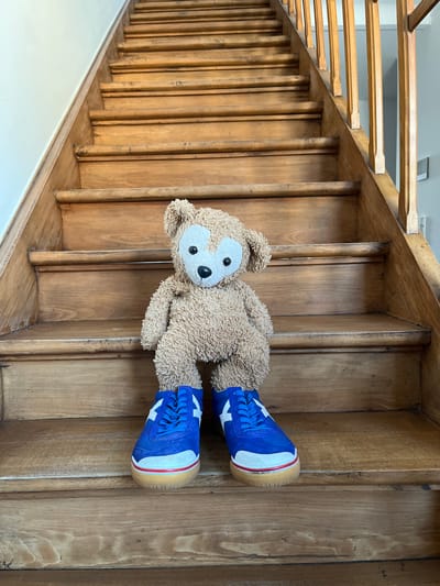 A light brown plush teddy bear is centrally positioned on the third wooden step of a staircase in a residential setting in Foetz, Luxembourg. The bear, featuring a lighter facial area and black eyes, has its head tilted slightly to the viewer's right. It is equipped with a pair of blue low-top athletic shoes, each displaying a white star motif on the lateral side, white toe caps, a white midsole accented with a red stripe, and a natural gum-colored outsole. The staircase consists of natural wood treads and risers, exhibiting varying degrees of wear and darkening, particularly at the edges. A pale-toned wall runs adjacent to the left side of the stairs, while a wooden spindle banister is visible on the right, beyond which lies another light-colored vertical surface.