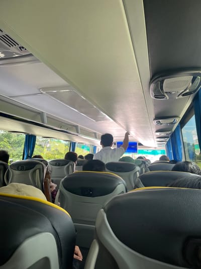 The image shows the interior of a bus, viewed from the perspective of a passenger seated towards the rear. The bus is filled with passengers, most of whom are seated and facing forward. The seating arrangement consists of rows of gray and black seats with yellow trim. Several passengers are visible, including a person in a white shirt in the aisle reaching up towards the ceiling, possibly adjusting an overhead compartment or a light. Outside the windows, lush green trees and vegetation are visible, suggesting a location in a tropical or semi-tropical environment. Blue curtains are drawn partially across the windows. The ceiling of the bus features air conditioning vents and overhead lights. The scene is typical of public transportation in the Philippines, likely on a route servicing or within the city of Koronadal, known for its agricultural landscape and warm climate.