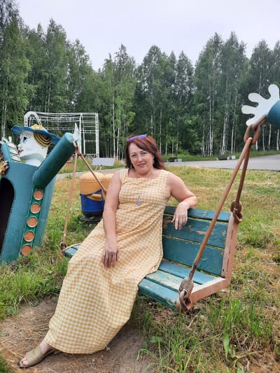 A woman with auburn hair, wearing a yellow and white checkered dress and gold sandals, is sitting on a weathered blue and brown swing. She is smiling at the camera, with her left arm resting on the back of the swing. Her right arm is bent, with her hand resting on her leg. She is wearing a necklace with a purple pendant and sunglasses are perched on her head. To her left, partially visible, is a large, teal-colored cartoon-like figure with a blue hat and yellow trim. The swing and figure are in a grassy area with scattered dry leaves. In the background, a line of birch trees forms a dense forest. A white metal structure, possibly part of a playground or carnival ride, is visible behind the cartoon figure. The scene appears to be outdoors in an amusement or park setting, possibly in the city of Zelenogorsky, Russia, given the prevalence of birch forests in the region.