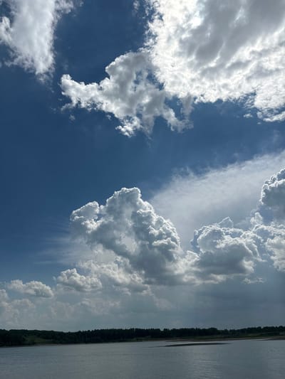 A wide-angle view captures a body of water, likely a river or lake, in the foreground, with a dark line of trees forming a natural horizon in the distance. Above the treeline, a vast expanse of sky dominates the scene. The sky is filled with a dramatic arrangement of clouds. The upper portion of the sky is a deep, clear blue, punctuated by large, puffy white clouds that appear to be illuminated by sunlight from above and to the right. Lower down, closer to the horizon, the clouds become denser and darker, suggesting the potential for rain or a storm. The overall impression is one of a powerful sky over a serene, natural landscape. There are no people or specific structures visible within the frame.