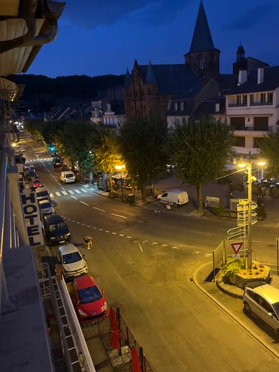 This is an aerial, long shot photograph taken at dusk in Espalion, France. The scene depicts a street lined with trees and buildings, with a prominent church spire in the background. Cars are parked along the street and a few are in motion. A person is bent down in the middle of the street, and another person is walking in the distance. The lighting suggests it is twilight, with streetlights and building lights illuminating the scene. A "HOTEL" sign is visible on the left side of the image.
